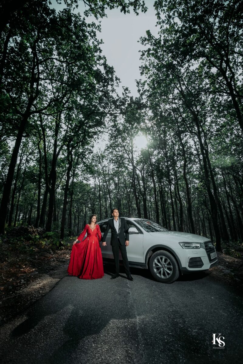 A couple poses in a dense forest, standing on a winding road next to a white SUV. The woman, in a vibrant red gown, holds the man's hand. He is dressed in a black suit. Tall, slender trees with dark trunks and green canopies dominate the background, with sunlight peeking through the leaves. The setting evokes a romantic and adventurous mood