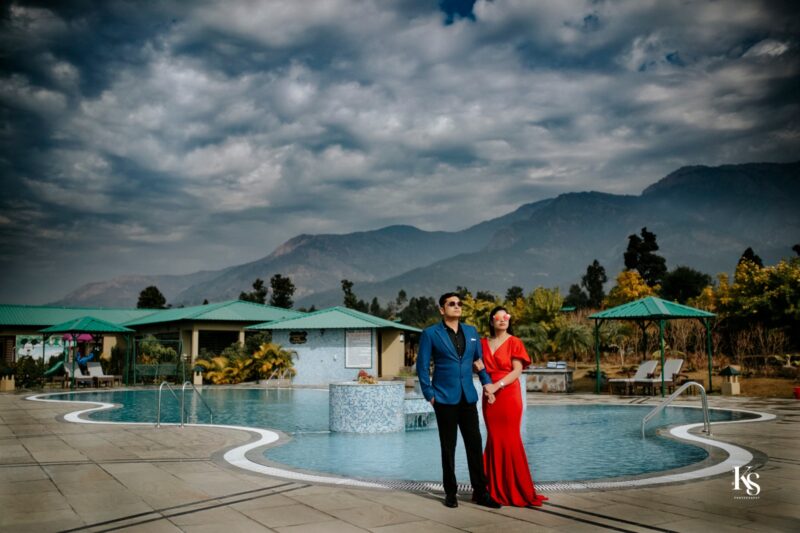 A couple stands beside a curved swimming pool with a tiled fountain in the center. The woman wears a long red dress, and the man is in a blue suit. Behind them are resort buildings with green roofs and lush trees, set against a backdrop of distant mountains under a dramatic, cloudy sky. This picturesque scene captures the essence of best pre wedding photography in Jim Corbett.